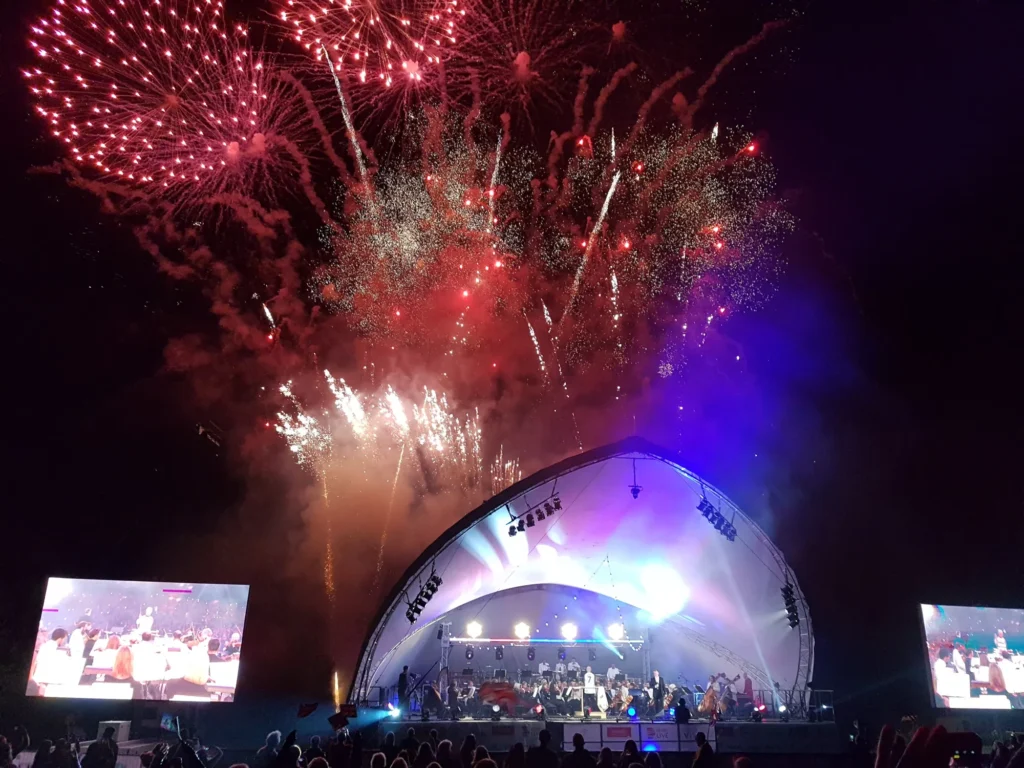 View from the audience of the Sinfonia Viva orchestra performing their annual Darley Park concert in Derby with fireworks going off over the stage, two large video screens (one on each side). The stage is lit up brightly and the full orchestra can be seen. On the far right hand side at the front of the stage, monitor mix engineer Jamie Rhodes-Simpson can just about be made out.