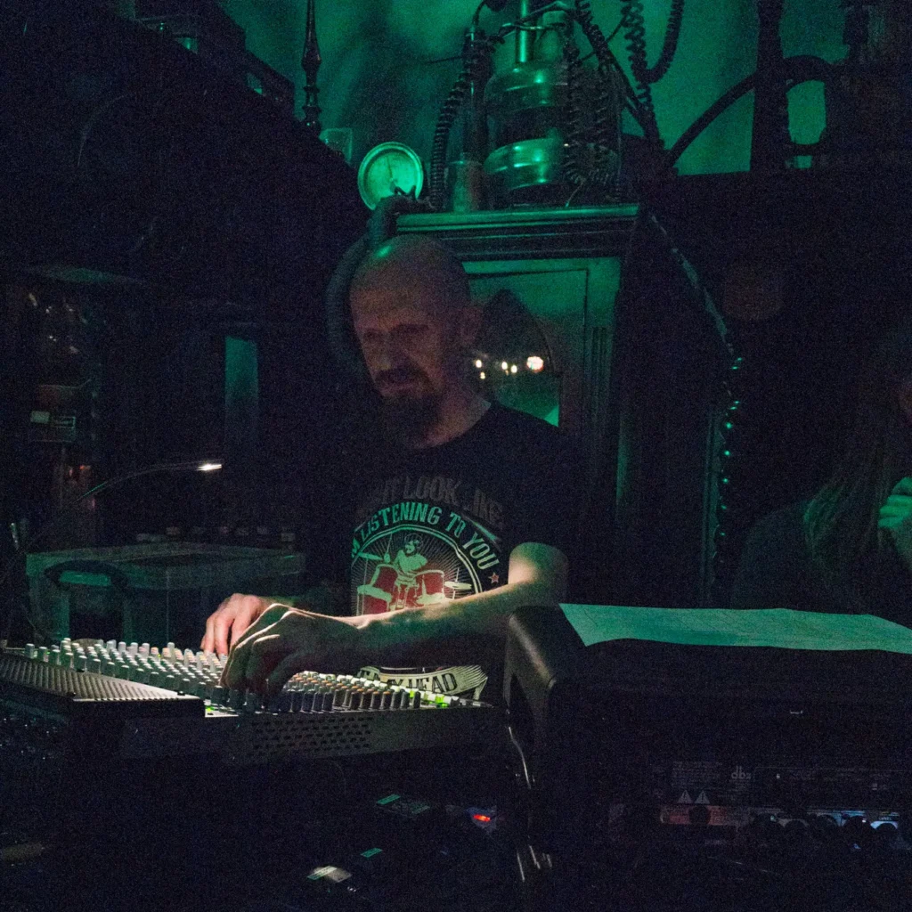 A very grainy photo of Sound engineer Jamie Rhodes-Simpson operating our Mackie 1640i analog mixing console at the Pit and Pendulum, Nottingham. To the right can be seen the rear of a rack of analog equipment (this included dbx compressors and Drawmer gates). Spooky apparatus like something out of Frankenstein can be seen lit up green high up behind Jamie.