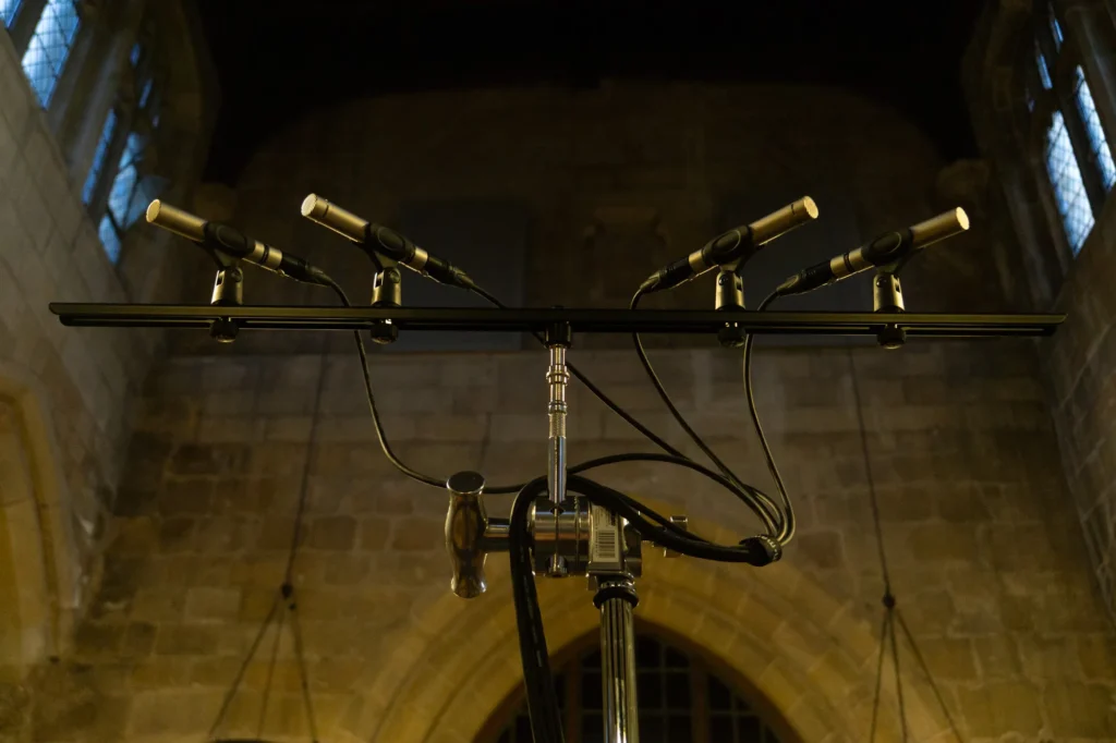 Image of four small diaphragm pencil condenser microphones on a bar high up in a church configured with two cardiods and two omni outriggers for recording a choir. The church stone walls and high up windows can be seen in the background lit from below.