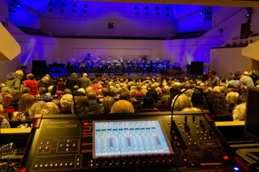 A full house (over 2200 people) present for the New Year’s Eve Sinfonia Viva concert at the Royal Concert Hall in Nottingham. Photo taken from the front of house mixing position just before the show starts. The stage is subtly lit with moody blue lighting. The orchestra music stands can be seen on the stage, the audience are still coming in, though most seats are already taken by this point. In the foreground a DiGiCo SD9 mixing console screen can be seen.