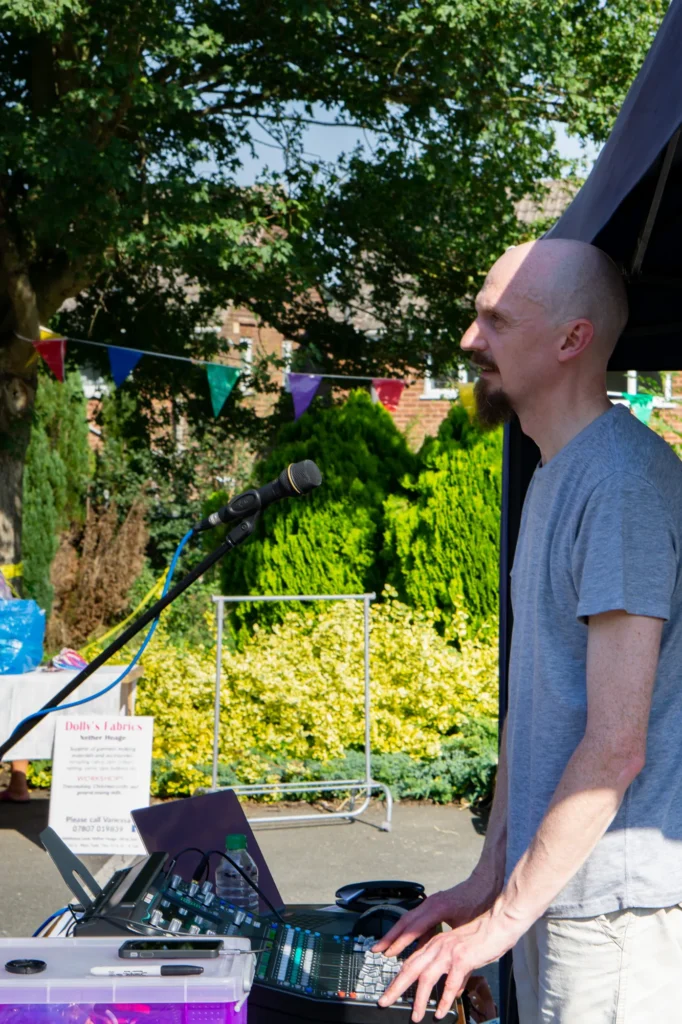 Sound engineer Jamie Rhodes-Simpson operating our Allen & Heath SQ-5 digital console at a charity event held at The Spanker Inn, Nether Heage, near Belper with Dave Kaye and the Dykons on stage (not shown in photo). Triangle flags on a string can be seen in the background along with green trees and bushes. It's a very bright and hot summers day.