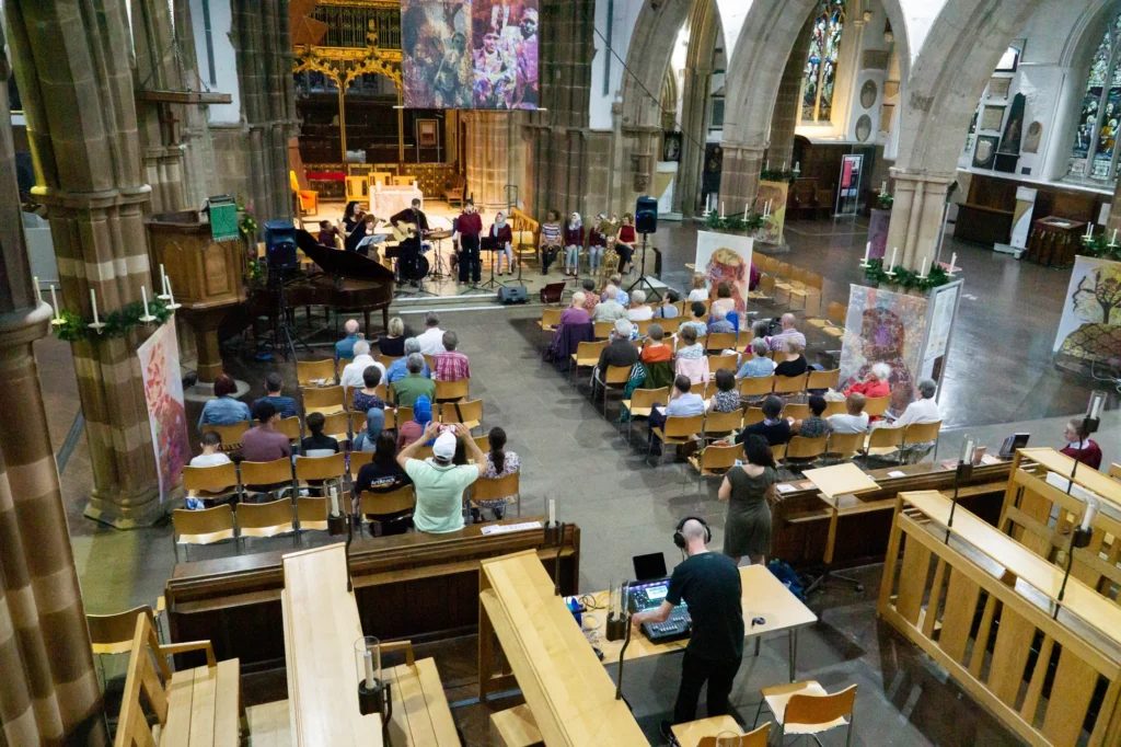 Wide angle photo taken from the balcony in Leicester Cathedral showing the Sanctuary Seekers Choir performing their Stories of Sanctuary concert at Leicester Cathedral. A grand piano, violinist, guitar player, drums, and various singers can be seen on stage in front of a gathered audience, with sound engineer Jamie Rhodes-Simpson mixing the show (and monitors) from behind the audience.