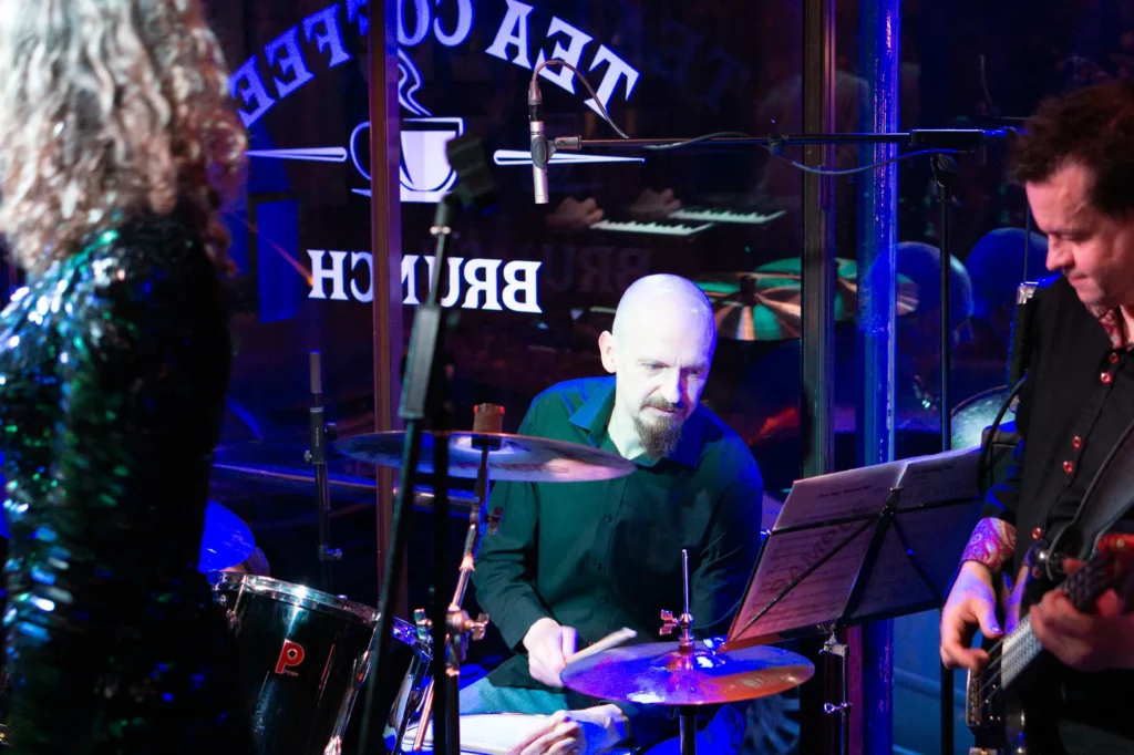 Drummer Jamie Rhodes-Simpson behind his Premier drum kit with Paiste cymbals performing on stage with Detroit Live Motown Tribute Band at Harrogate Blues Bar. Singer Anna Shotter can be seen on the left of the image, and bassist Graham Alexander on the right. The stage is lit up in blue light. Two small diaphragm condensor mics in a Glyn Johns style set up can be seen overhead and behind the drums.