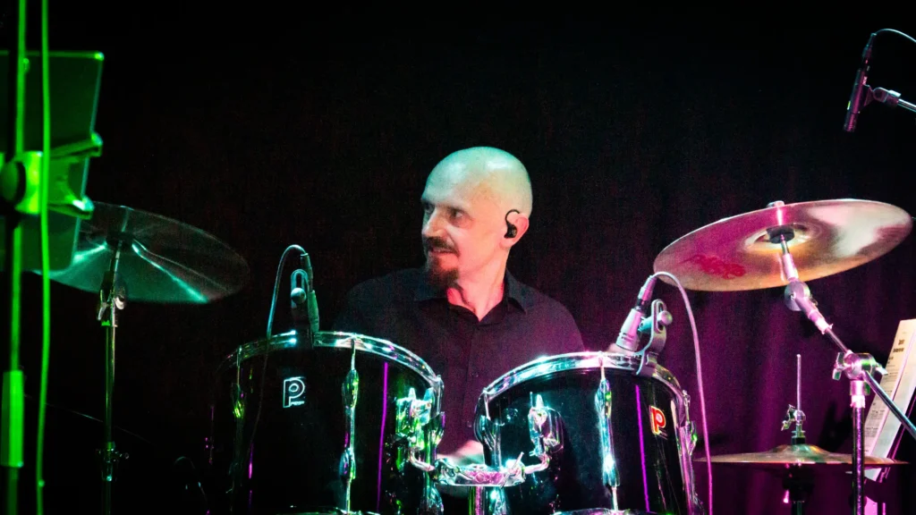 Drummer Jamie Rhodes-Simpson wearing IEMs behind his Premier drum kit with Paiste cymbals performing on stage with Detroit Live Motown Tribute Band at the Old Courts in Wigan. On the left stage lights colour a stand green, whilst the right hand side of the image purple, which is also highlighting the black drum shells.