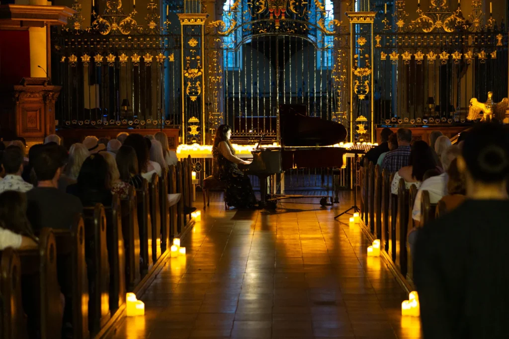 A pianist plays by candlelight at Derby Cathedral. Rows of wooden pews can be seen with candles by their side. Hundreds of candles are also on tables in the performance area. The gold on the grand iron railings glowing elegantly in the candlelight. The front of house sound engineer is Jamie Rhodes-Simpson (not seen in photo).