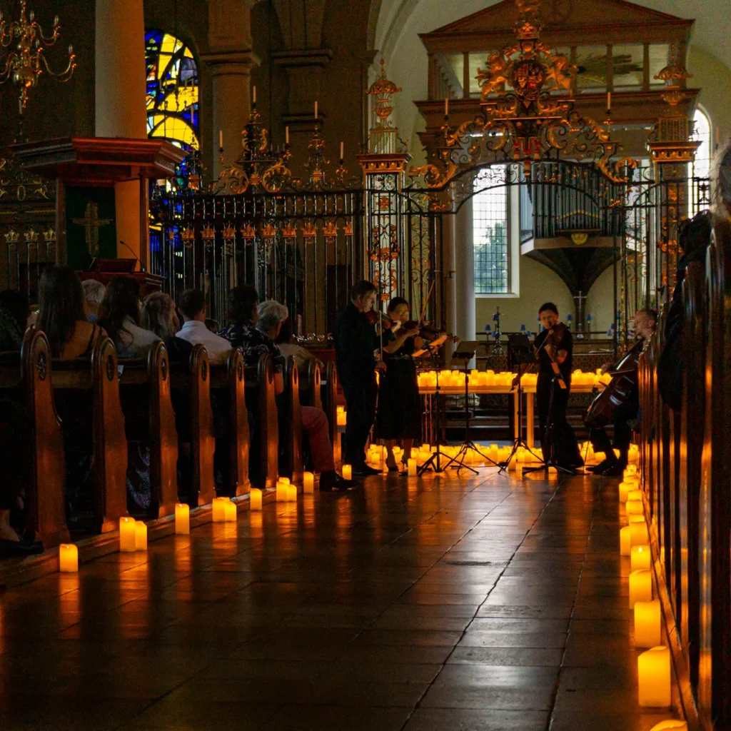 A string quartet plays by candlelight at Derby Cathedral. Wooden pews can be seen with candles at the end of each row. Hundreds of candles are also on tables in the performance area. The gold on the grand iron railings glowing in the candlelight. Behind the railings can be seen a stained glass window with blue and yellow panes of glass lit up with the last vestiges of daylight, and also pipes from one of the pipe organs. The front of house sound engineer is Jamie Rhodes-Simpson (not seen in photo).