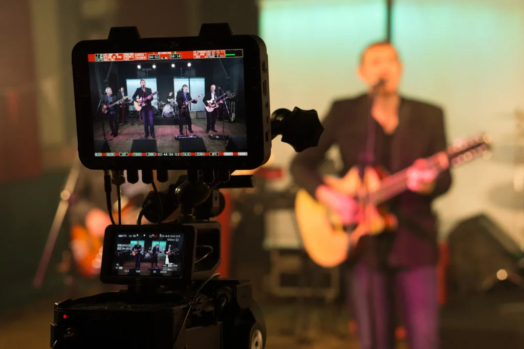 Jamie Rhodes-Simpson (not shown) filming a promo video for UK Eagles tribute band Motel California, who are out of focus in the photo (shallow depth of field) performing on a set in the background. In the foreground a Sony FS700 camera has an Odyssey 7Q+ recorder mounted above it with the band clearly visible on the screen.