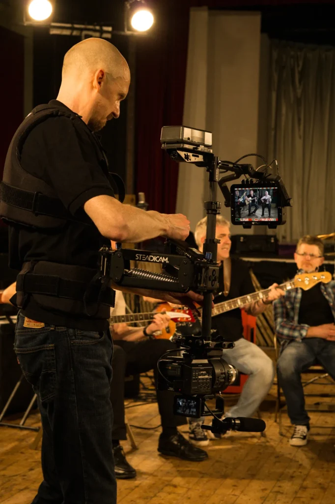 Jamie Rhodes-Simpson operating his Steadicam rig in low mode (upside down) configured with a Sony FS700 camera filming a promo video for UK Eagles tribute band Motel California, who are performing a song sat down in the background of the photo. Stage lighting can be seen high up on the left and a stage in the background.