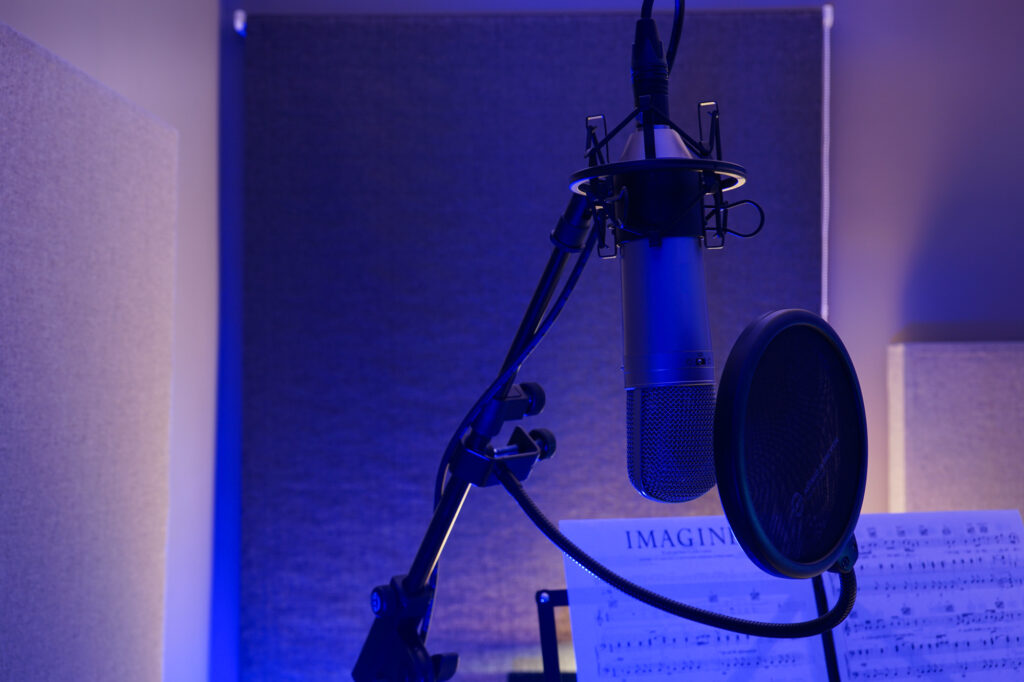 Close up shot with mood lighting of a large diaphragm multi-pattern condenser microphone hanging upside down in a suspension basket with a pop filter and sheet music for John Lennon's Image on a music stand. The corner of a sound absorption panel can be seen on the left of the image.