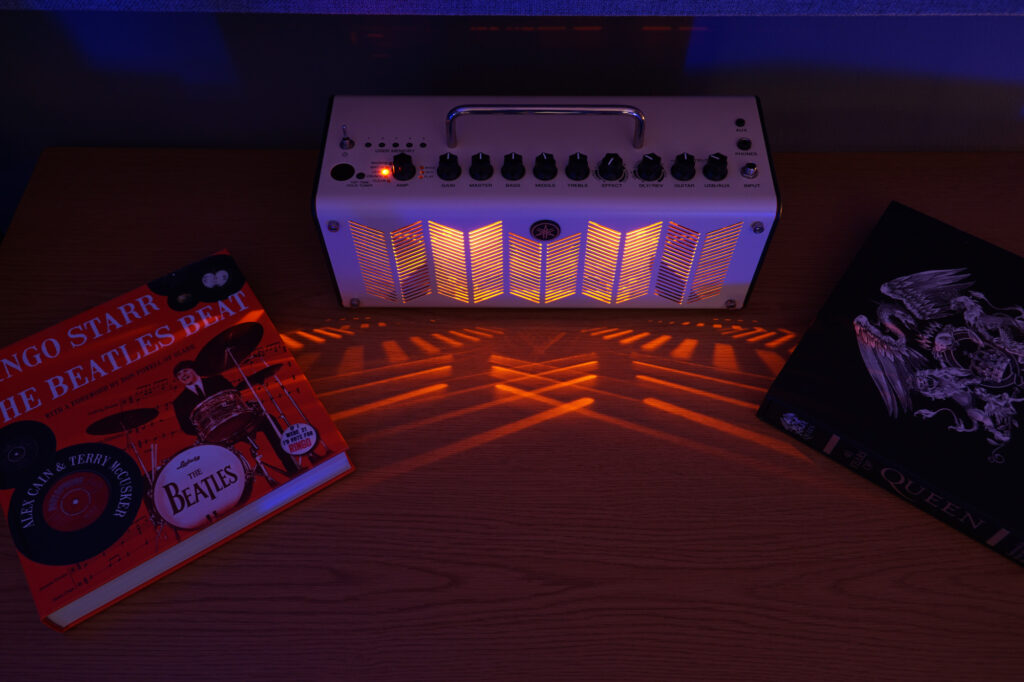 Photograph of our Yamaha THR10 guitar amplifier emitting a warm tube like glow casting orange light and dark shadows onto a table with books about Beatles drummer Ringo Starr and Queen.