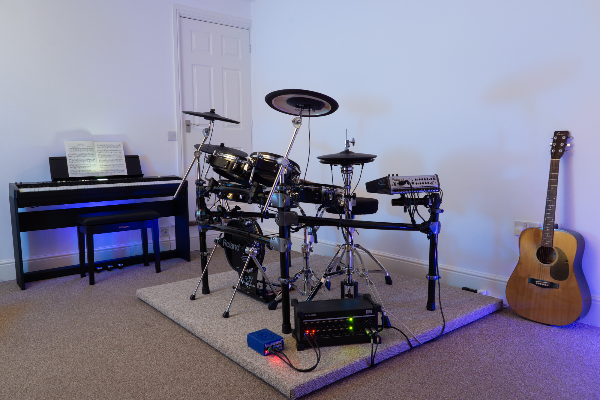 Photo of the Capybara Sound studio live room showing Roland TD-20 drums on a custom built drum riser, Roland FP-E50 digital piano, an acoustic guitar, Allen & Heath DX168 extender/stage box and a Klark-Teknik DN200 DI box.