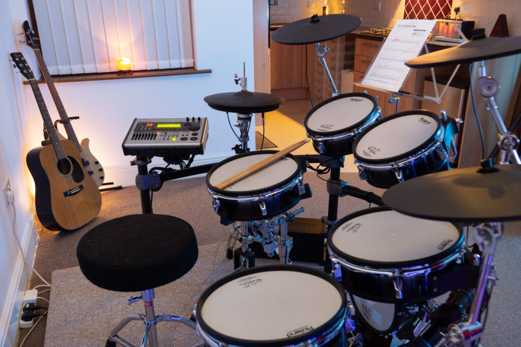 Photograph of the Roland TD-20 drum kit in the Capybara Sound studio live room with an acoustic guitar and Yamaha bass in the near background, and a warm glow from a kitchen area seen in the distance. A salt lamp glows pleasantly on the windowsill behind the guitars.