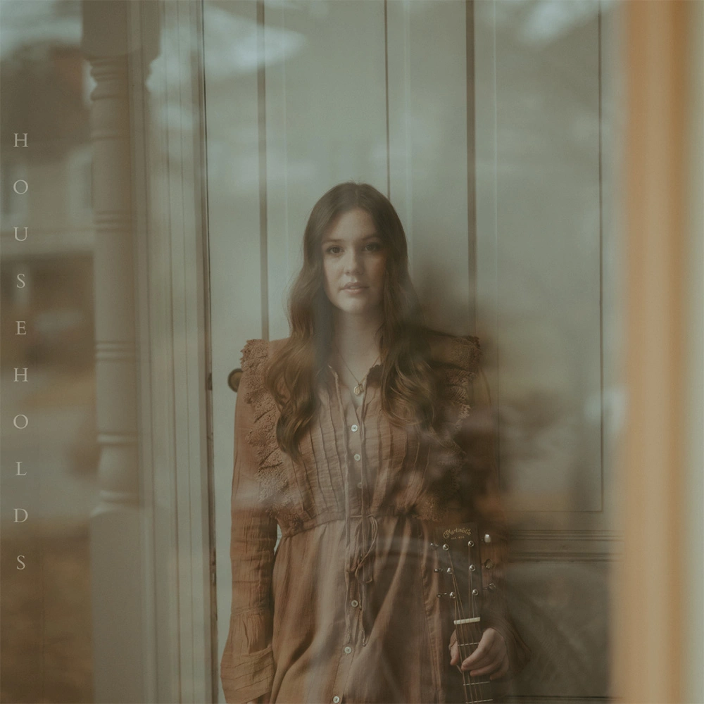 Cover art for Households song by Gloria Anderson. Gloria is shown standing holding the neck of a guitar (only the top part of the fretboard is visible). The image appears as if seen through a window with reflections in the glass.