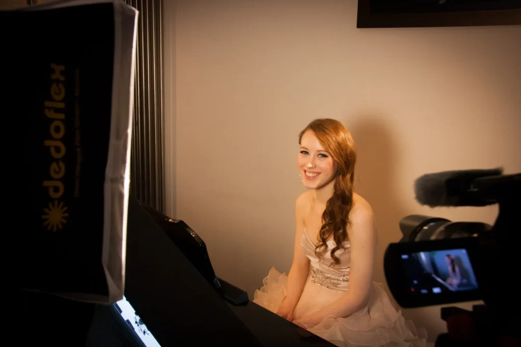 Behind the scenes photo from a video shoot with singer Hannah-Rei. A Dedo soft light can be seen to the left with Hannah lit in the centre wearing a glamourous dress sat behind a piano. To the right the video camera is partially in shot. The photo has a nice warm feel to it with a golden glow to the lighting. Filmed and lit by Jamie Rhodes-Simpson (not seen in photo).