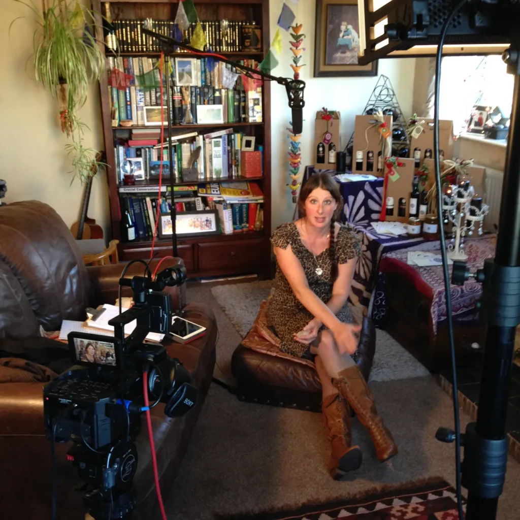 Video producer and sound engineer Jamie Rhodes-Simpson (not shown) filming Karen Heyhoe for an Aradia promo video. The photo shows Karen in the centre, lit by a studio light just visible on a stand in the foreground. The the left is a Sony FS700 camera wired to a shotgun condensor mic on a stand above Karen's head. On the back wall is a bookcase with a large spider plant to the left, and a display of Aradia products (fine wines, olive oil and honey) on the right.