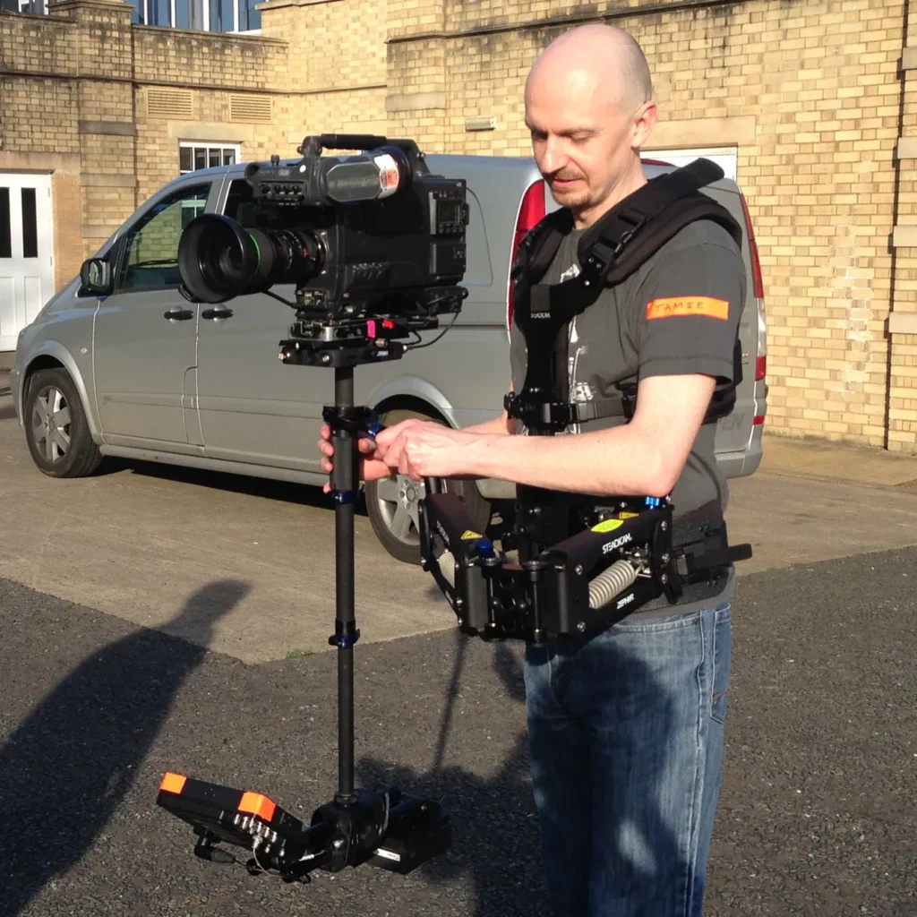 Jamie Rhodes-Simpson operating a Steadicam with a full sized Sony television camera mounted. There is a silver van in the background parked next to a beige brick building.