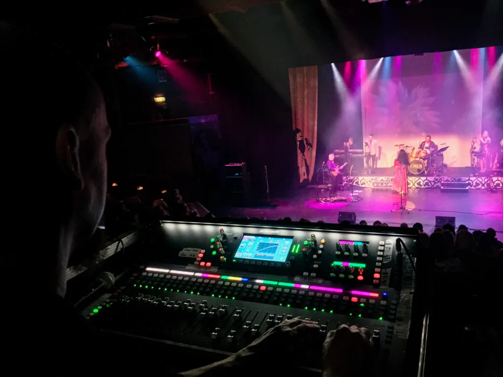 Cher tribute band performing on the large stage at the Medina Theatre, Newport, Isle of Wight. Sound engineer Jamie Rhodes-Simpson mixing front of house sound (and monitors) using an Allen & Heath SQ-7 digital console. Stage lights can been seen streaming from overhead, blue, purple, red and white colours. On stage are singers, guitarists, keyboards, drums and other musicians. A full audience can just be made out in the darkness between front of house and the stage.