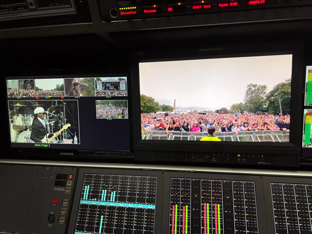 Video monitors above the Calrec sound console in the TV outside broadcast truck for King Tuts stage at Glasgow TRNSMT Festival. One monitor is showing a large crowd waiting in front of the King Tuts stage amd another is showing various camera angles and Nile Rodgers & Chic performing on the main stage.