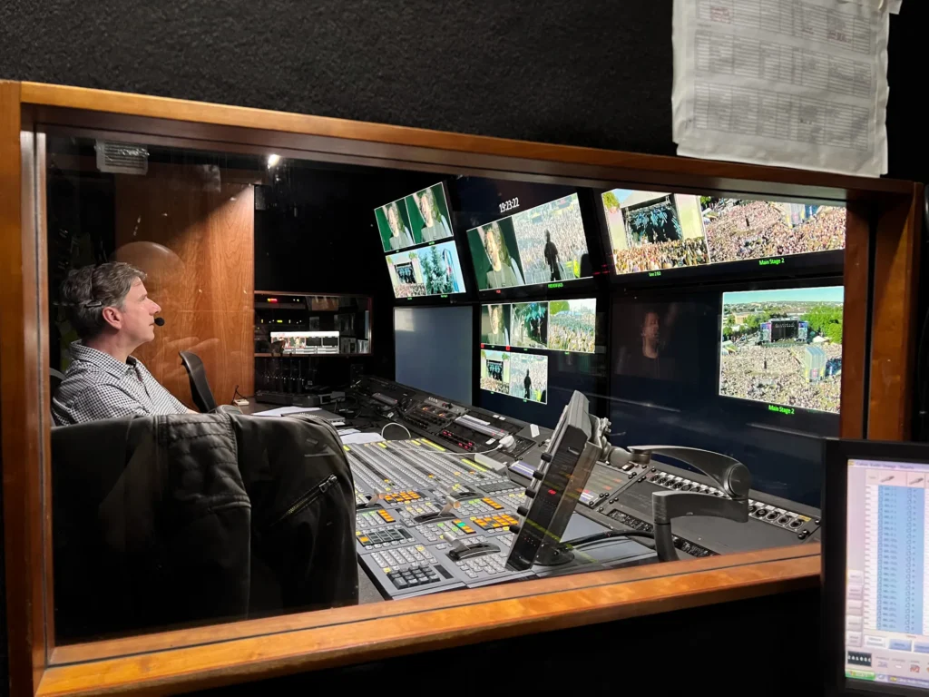 View of TV outside broadcast truck gallery showing Donald MacInnes Directing and Vision Mixing TRNSMT Festival, King Tuts Stage, Glasgow. Lots of monitors cover an entire wall showing different camera angles. A large vision mixer board plus various comms panels are also seen through a window from the sound area of the truck.