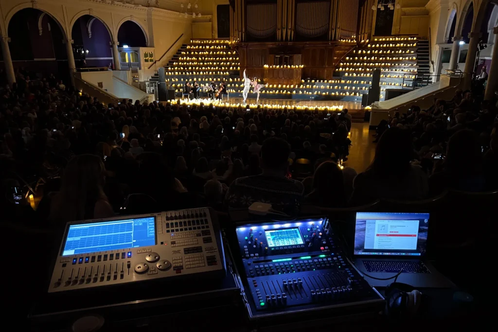 View from front of house mix position for a Candle Light Concert Ballet. An Avolites lighting control desk, Allen & Heath SQ-5 digital mixing console and a MacBook Pro computer are shown. Hundreds of candles can be seen on the choir steps up each side of the stage with a large pipe organ central. In front on the stage is a string quartet and a ballet couple dancing. Front of house sound by engineer Jamie Rhodes-Simpson (not shown).