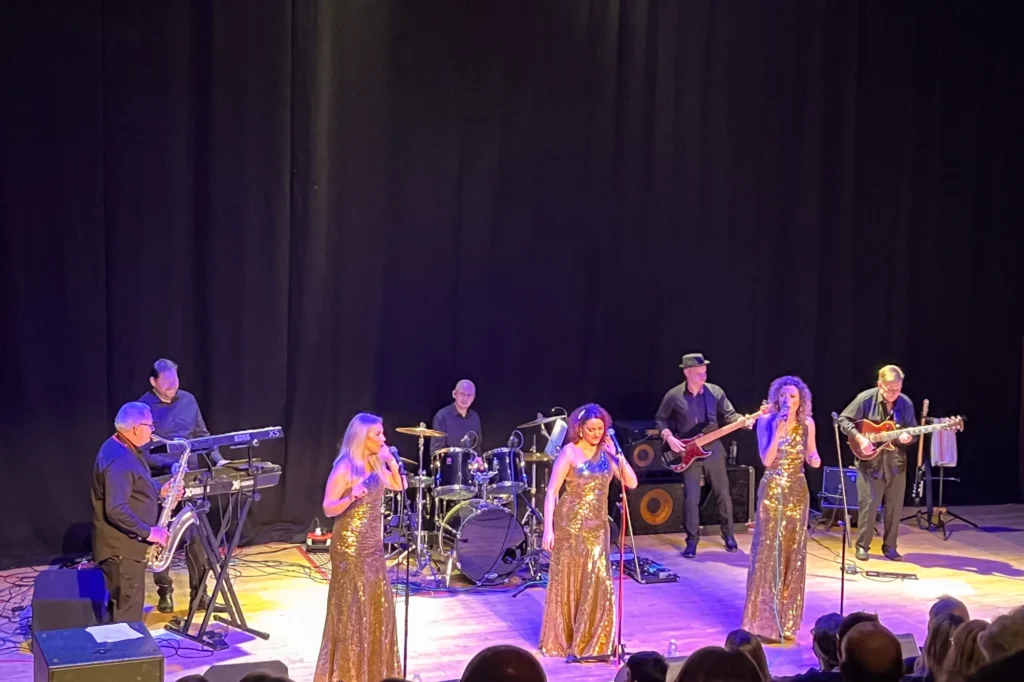 Detroit Live Motown Tribute Band performing on the sizeable stage in the Red Brick Auditorium theatre at Square Chapel Arts in Halifax UK. From left to right, Mark Witty (sax), Wayne Mitchell (keyboards), Clare Campbell (vocals), Jamie Rhodes-Simpson (drums), Hayley Arnold )(vocals), Graham Alexander (bass), Anna Shotter (vocals), Chuck Annan (guitar). Glamorous gold full length dresses for the three female singers and the band all dressed in black shirts and trousers. A few rows of audience can just be seen at the bottom of the picture.