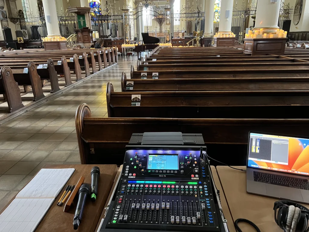 Two Sennheiser hand held radio mics, an Allen & Heath SQ-5 digital mixing console and a MacBook Pro computer displaying Sennheiser WSM radio mic and IEM control software can be seen setup behind row after row of wooden pews in Derby Cathedral. In the distance a grand piano can be seen with the lid open. Candles have been placed around the building ahead of a Candlelight Concert later. The grand metal railings can be seen that separate the cathedral into two distinct areas. The front of house sound engineer is Jamie Rhodes-Simpson (not seen in photo).