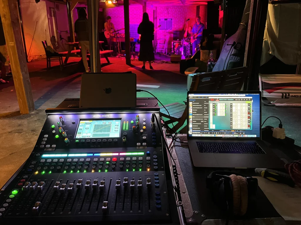 Newark festival. Local band at night, lit by purple and red lighting. An Allen & Heath SQ-5 digital mixing console and Apple MacBook Pro laptop are shown in the foreground. The laptop showing controls for dbx VENU360 PA system processors. Moody but warm and inviting night time shot. Sound engineer (not shown) is Jamie Rhodes-Simpson on front of house mixing (monitors from FoH).