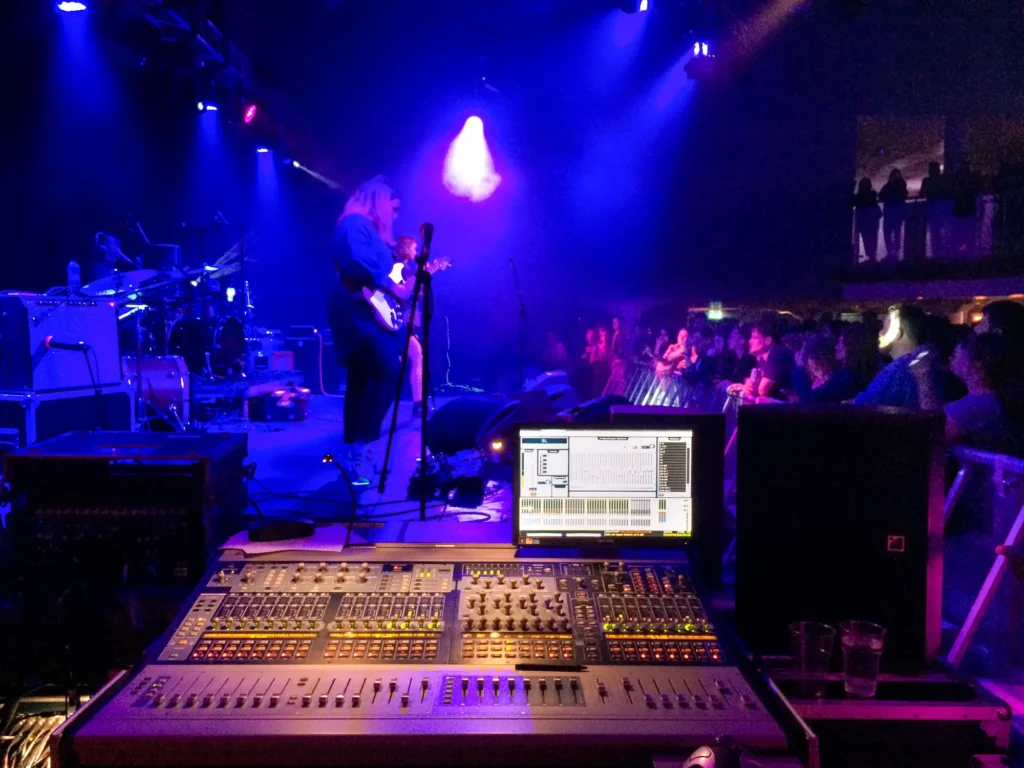 View from side of stage monitor position (sound engineer Jamie Rhodes-Simpson operating - not shown). An Avid VENUE Profile mixing console can be seen with the control screen and an L-Acoustics monitor speaker to one side. The stage is lit up in blue from the moving lights and the band "Swimming Girls" can be seen performing at the Dot to Dot Festival in Nottingham. The venue is Nottingham Trent University Student Union. A crowd barrier just in front of the stage holds back an eager audience. More people can be seen on the balcony in the background.