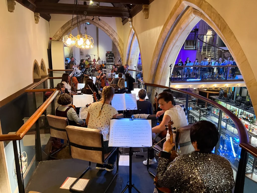 UFO Orchestra rehearsing at the Pitcher & Piano in Nottingham, for the Hockley Hustle, Festival. A large old church building saw the orchestra positioned around the balcony area. The photo shows the string section, with part of the woodwinds visible on the balcony bridge. Violins, violas and cellos, then drums and percussion in the background and furthest away, double bass players. Many microphones are set up, including spot mics on various instruments. The amplifier rack, radio mics and other audio I/O can be seen stacked in a tower in the distance.