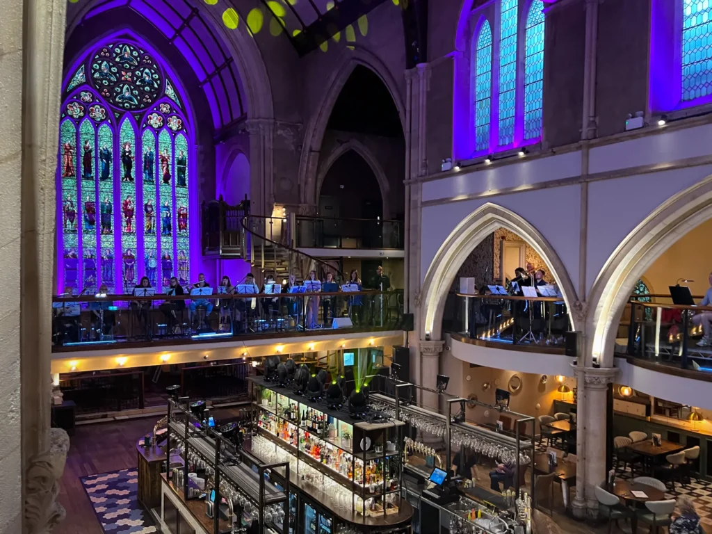 UFO Orchestra concert at the Pitcher & Piano in Nottingham, for the Hockley Hustle, Festival. A large old church building with tall walls lit up in purples and reds and laser lights beaming out from the balcony. The orchestra are all around the balcony area (strings & percussion on the left - not visible in photo, woodwinds across the bridge and brass on the right). Photo taken during rehearsals, so the bar area is empty. A very large and colourful stained glass wind is seen to the rear of the building.