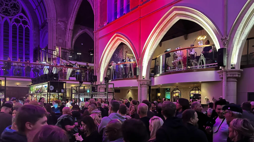 UFO Orchestra concert at the Pitcher & Piano in Nottingham, for the Hockley Hustle, Festival. A large old church building with tall walls lit up in purples and reds and laser lights beaming out from the balcony. The orchestra are all around the balcony area (strings, percussion on the left - not in photo, woodwinds across the bridge and brass to the right). A large crowd is in the main bar area on the ground floor.