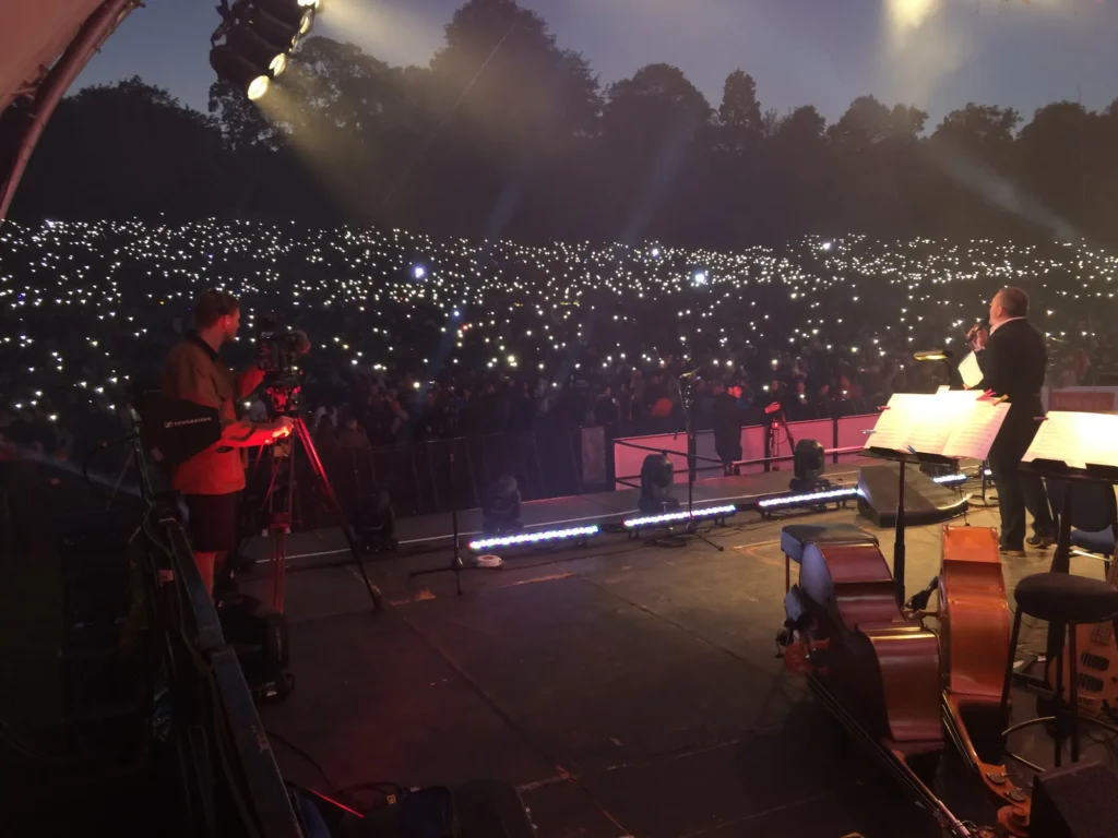 View from on stage at the Darley Park Classical Concert in Derby. Dusk is well advanced and thousands of mobile phone lights can be seen in the audience. On stage a presenter is talking and two double bass can be seen laid down in the foreground.