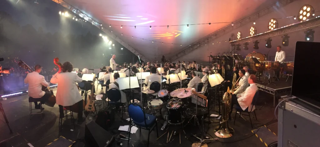 View from the monitor mix position of the full Sinfonia Viva orchestra performing at the Darley Park concert in Derby. Lights on each music stand, plus stage lighting and moving lights can be seen. The monitor sound engineer is Jamie Rhodes-Simpson (not shown in photo).