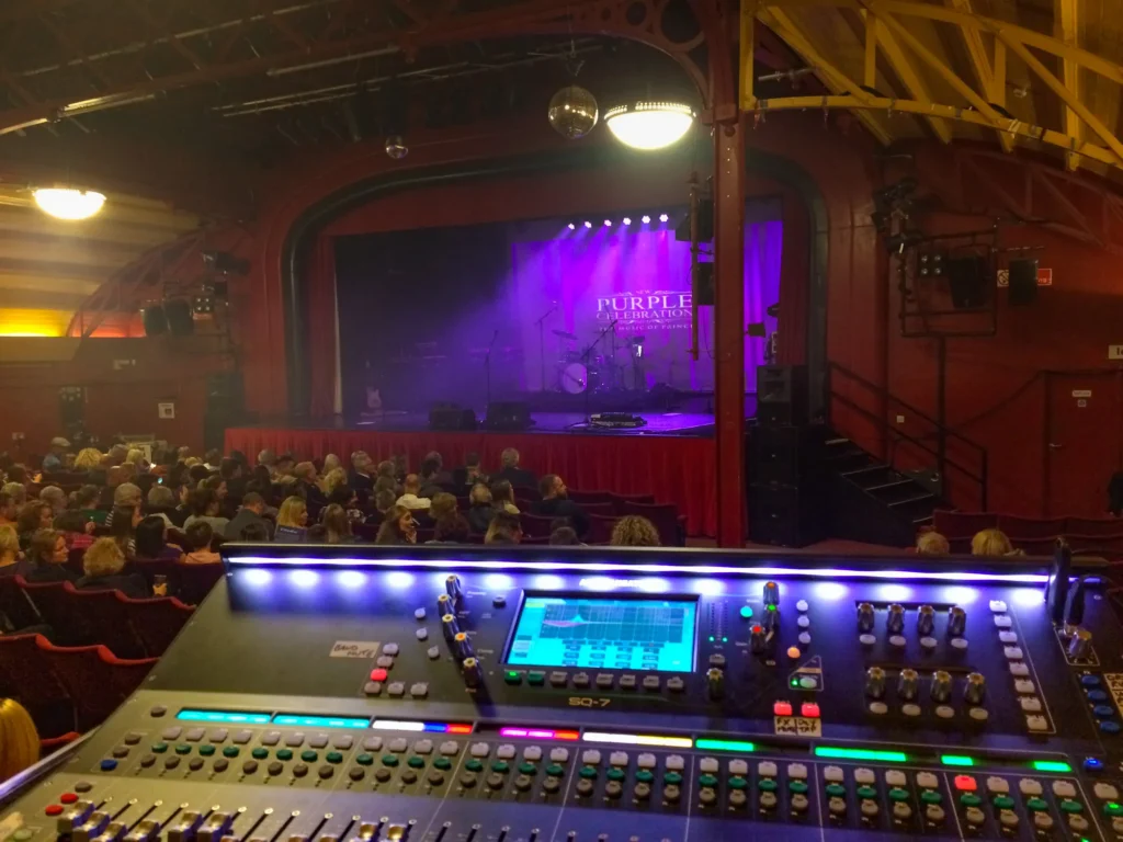 New Purple Celebration - the music of Prince. Sound engineer Jamie Rhodes-Simpson (not shown) behind an Allen & Heath SQ-7 digital mixing console mixing front of house sound. The audience is mostly in, only a few seats empty. Purple lights on the stage with the band name projected behind. Drums and keyboards can be seen. Anticipation before the start of the concert at The Pier Theatre in Cromer.