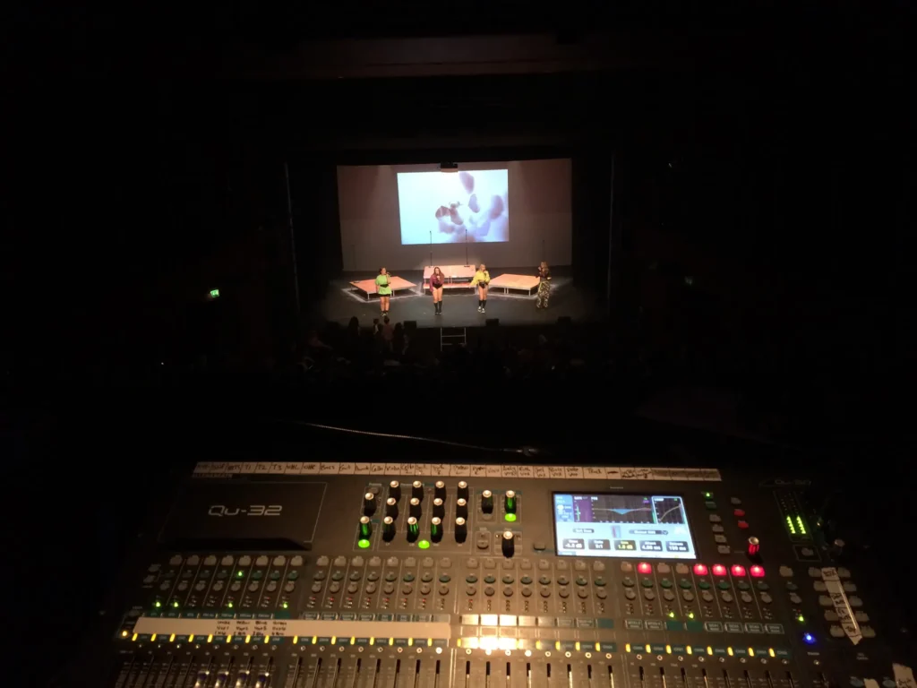 The Little Mix Experience girl group singing live on stage at the Lighthouse Theatre in Poole. Sound engineer Jamie Rhodes-Simpson (not shown) operating an Allen & Heath Qu-32 mixing console for front of house and IEM sound. The console is high up behind the audience, the stage looking quite small in the distance.