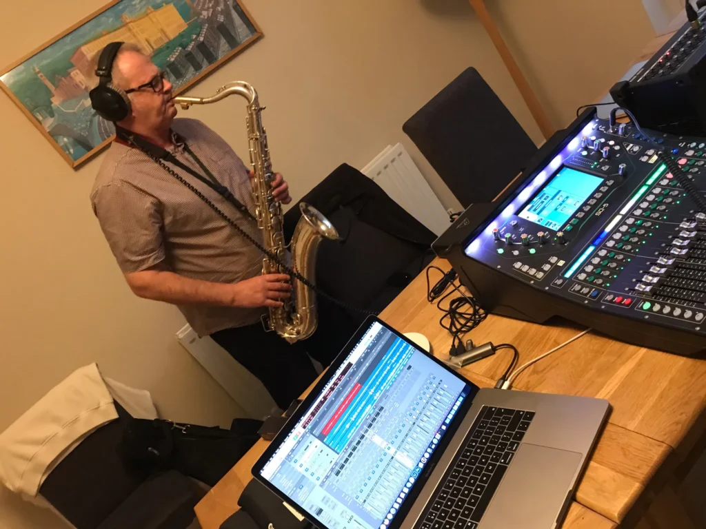 Saxophonist Mark Witty standing and playing tenor sax, overdubbing an earlier recording in his dining room at home. In the foreground on the table, our Allen & Heath SQ-5 mixing console, DX168 stage box and a MacBook Pro laptop running Logic. Mark is wearing beyerdynamic DT770-Pro headphones for monitoring purposes and to hear the playback. Recording engineer is Jamie Rhodes-Simpson (not seen in the photo).