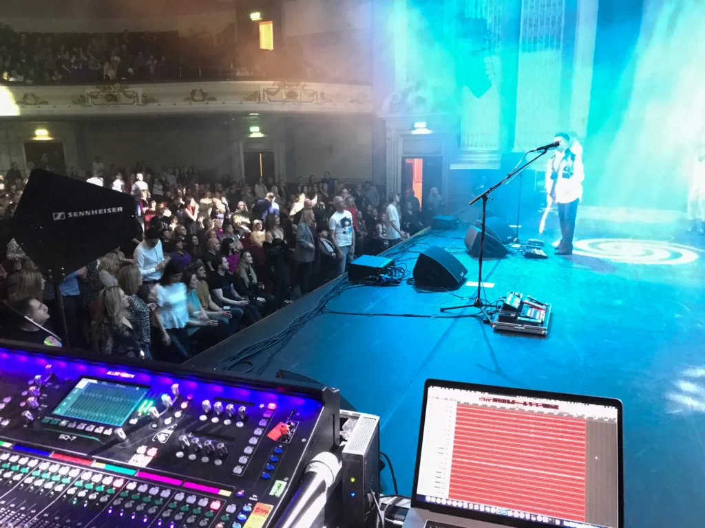 New Purple Celebration - the music of Prince perform at Usher Hall in Edinburgh. Viewed from side of stage at the monitor mixing position. A Sennheiser wireless antenna next to an Allen & Heath SQ-7 can be seen with an Apple MacBook Pro alongside recording the full multitrack output from the mixing console. Recording engineer is Jamie Rhodes-Simpson (not shown). Monitor mix engineer is Percy Young (also not seen in image). The band are on stage performing with cyan coloured moving lights (with gobos projecting onto the floor) streaming across the stage. The audience can be clearly seen with the amount of light coming off the stage and it's a very full house.