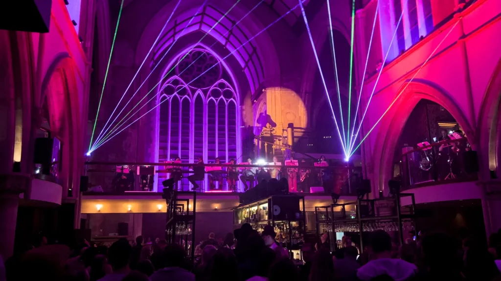 UFO Orchestra concert at the Pitcher & Piano in Nottingham, for the Hockley Hustle, Festival. A large old church building with tall walls lit up in purples and reds and laser lights beaming out from the balcony. The orchestra are all around the balcony area (strings, percussion on the left, woodwinds across the bridge and brass on the right). A guest singer is in the spotlight, high up on a spiral staircase. A large crowd is in the main bar area on the ground floor.